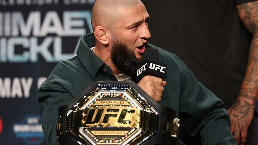 Khamzat Chimaev speaking into a UFC microphone at the UFC 328 Newark press conference, holding the Middleweight Championship belt